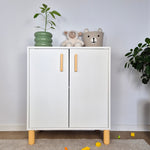 White cabinet with wooden legs and handles, topped with a plant and children's toys, against a light gray wall.