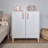 White cabinet with pink legs and doors in a room with toys and a lamp.