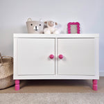 White cabinet with pink furniture legs, stuffed toys, and a decorative frame, showcasing stylish furniture legs.