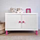 White cabinet with pink furniture legs, stuffed toys, and a decorative frame, showcasing stylish furniture legs.