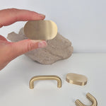 Hand holding a flat oval brass cabinet pull, showcasing three designs of brass cabinet pulls on a textured surface.