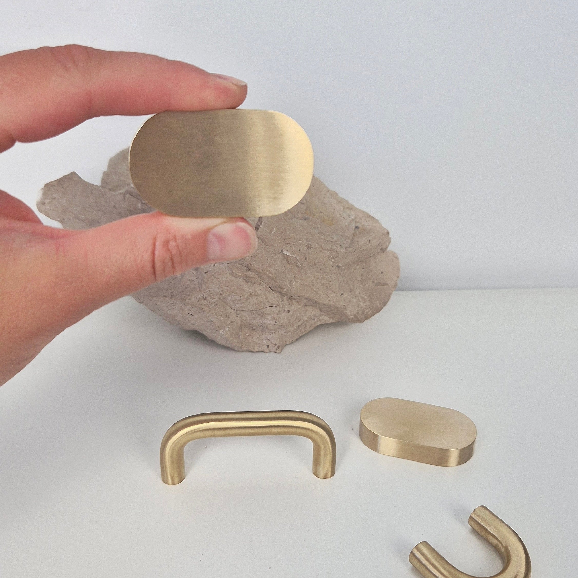 Hand holding a flat oval brass cabinet pull, showcasing three designs of brass cabinet pulls on a textured surface.