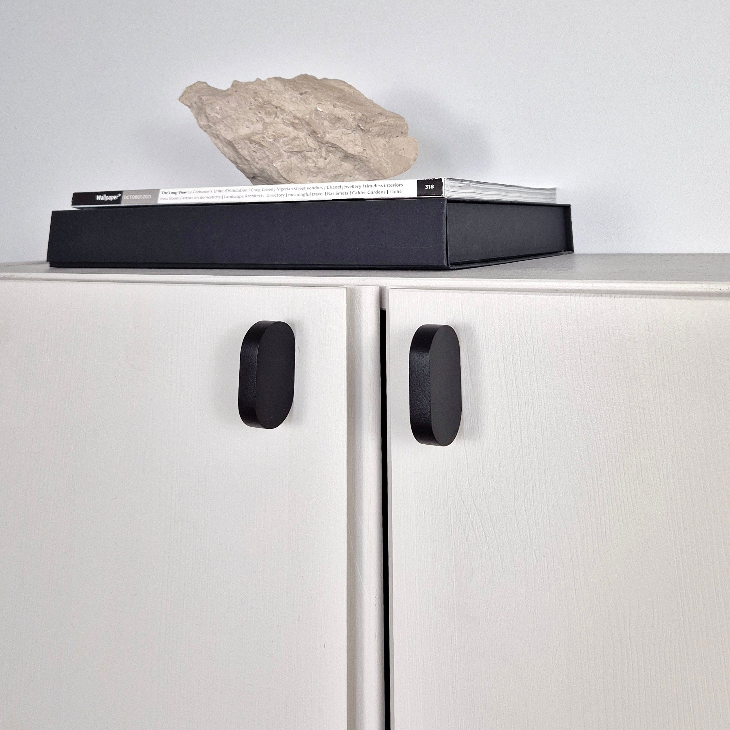 A modern white cabinet with two doors and matte black handles, topped with a black rectangular box and a light beige stone, against a plain light gray wall.