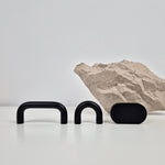 Three matte black hardware pieces, including two U-shaped handles and one oval knob, displayed on a white surface with a textured stone prop against a light gray background.