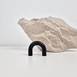 A matte black U-shaped metal hook displayed on a white surface in front of a textured light beige stone