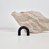 A matte black U-shaped metal hook displayed on a white surface in front of a textured light beige stone