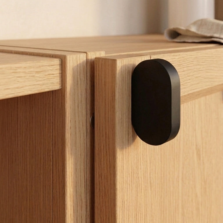 A close-up of a light oak wood cabinet featuring a matte black oval door pull handle, with a softly blurred kitchen background.